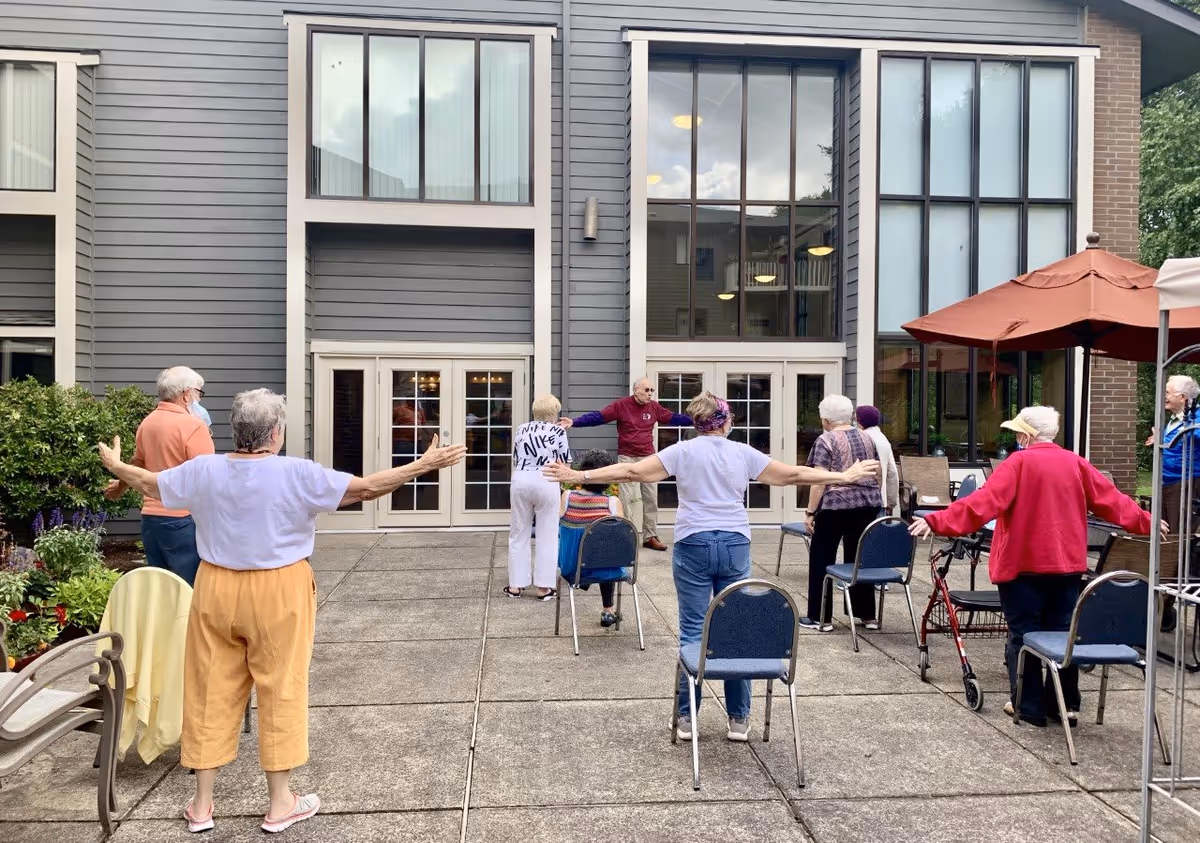 A group of elderly people participating in an outdoor exercise session on a patio area outside a building with large windows. They are standing with arms outstretched, some using chairs or walkers for support. The setting includes plants, patio furniture, and an umbrella.