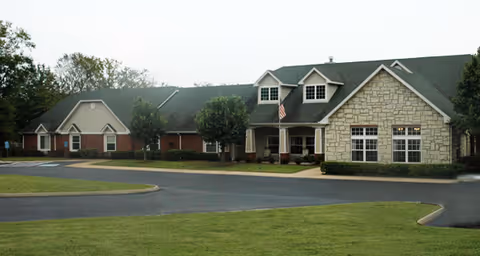 Single-story senior living facility with brick and stone facade, a covered entrance with an American flag, driveway and landscaped lawn.