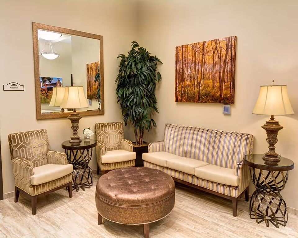 A cozy sitting area in an assisted living facility featuring a striped sofa, two patterned armchairs, a round brown leather ottoman, two side tables with decorative lamps, a large mirror on the wall, a tall green plant in the corner, and a framed picture of a forest with autumn trees.