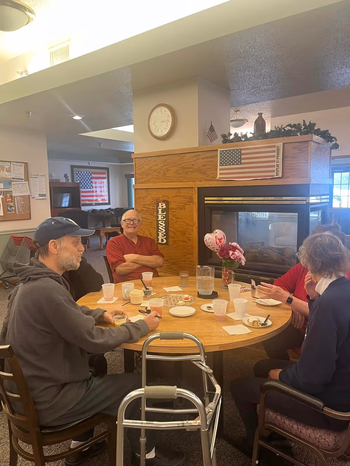 Several people sitting around a round table in a common room with cups and plates, a walker in front, and a fireplace and American flag in the background.