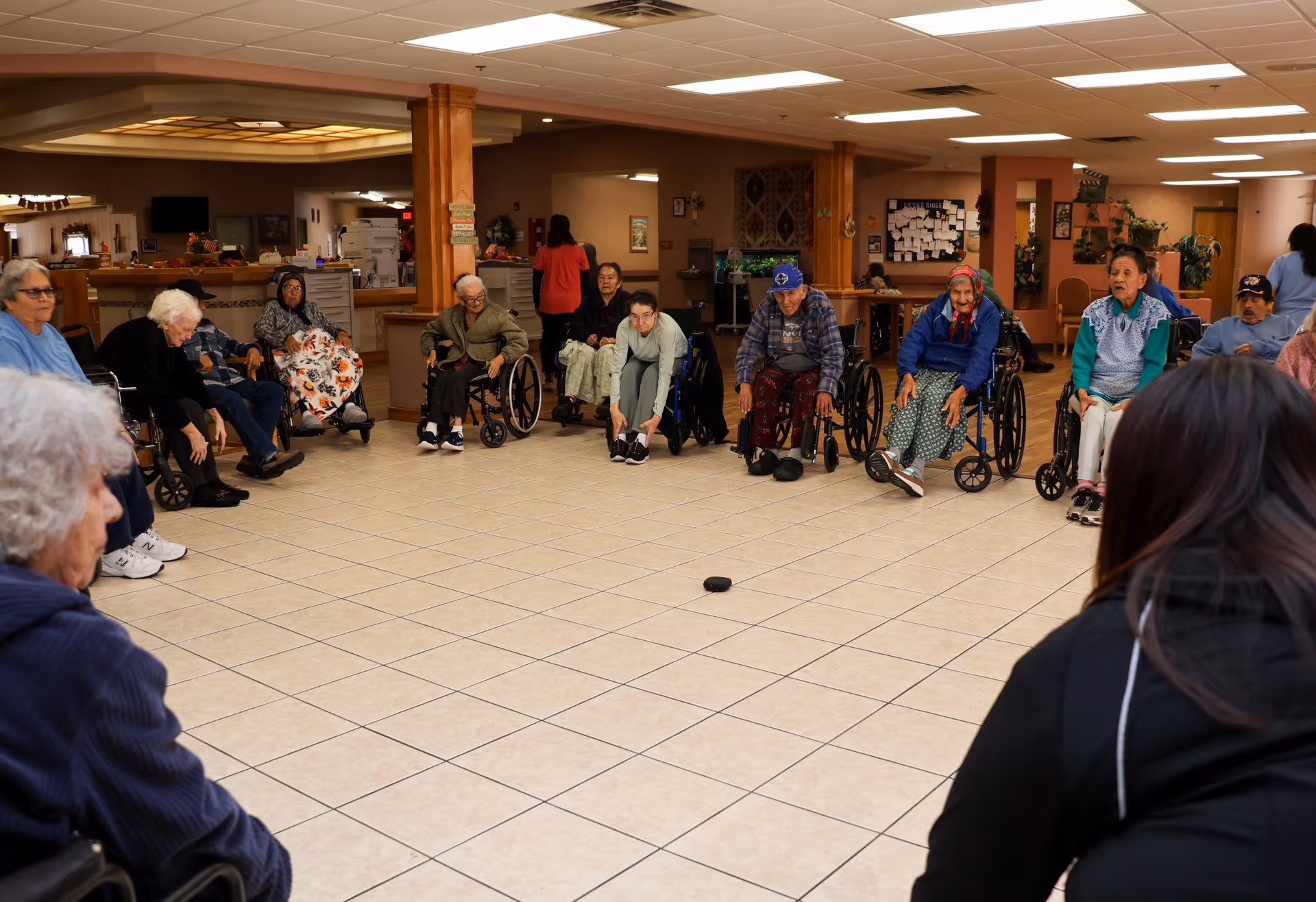 A group of elderly individuals, many in wheelchairs, seated in a large circle in a spacious room with tiled floors and wooden pillars, participating in a group activity. A staff member is seen in the foreground facing the group.
