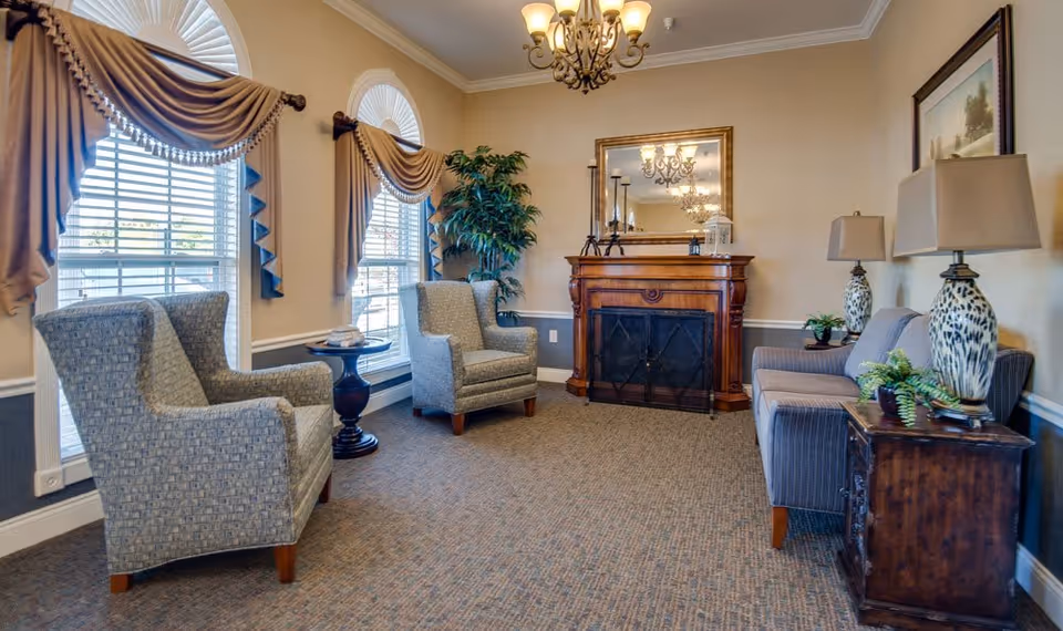 A cozy living room area with two patterned armchairs and a small round table between them, a wooden fireplace with a large mirror above it, a sofa with two decorative lamps on side tables, a potted plant in the corner, and windows with elegant draped curtains letting in natural light.