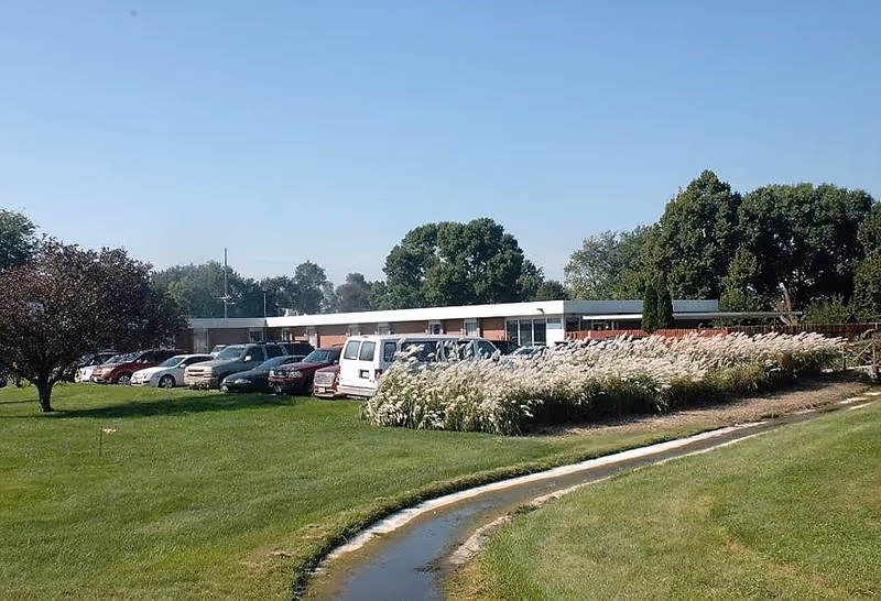 Low single-story senior living building with parked cars, ornamental grasses and a manicured lawn under a clear blue sky.