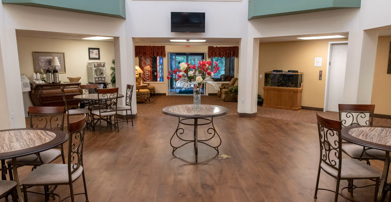 Interior of a senior living facility common area with round tables and chairs arranged on a wooden floor. A round table with a vase of red and white flowers is centered in the foreground. In the background, there is a seating area with armchairs, a Coca-Cola vending machine, a fish tank, and a wall-mounted TV above the entrance door.