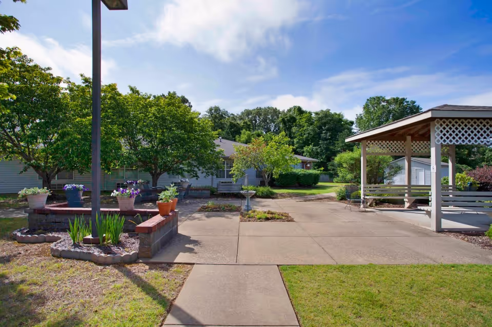 Outdoor area of Wellington Manor Jackson featuring a paved walkway, a small garden with potted plants and flowers, several trees, and a covered seating area with benches. The sky is partly cloudy and the surroundings are green and well-maintained.