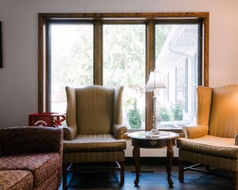 A cozy sitting area with two beige wingback chairs and a small round wooden table with a lamp between them, positioned in front of a large window with a view of greenery outside. A patterned sofa is partially visible on the left side.
