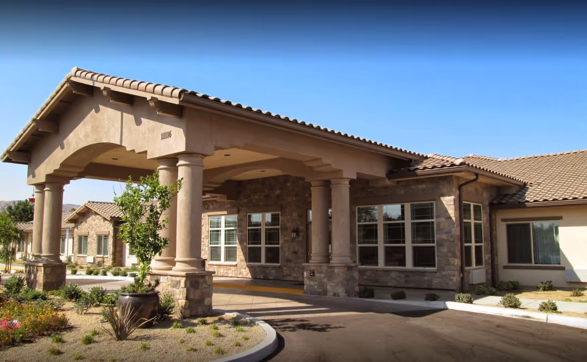 Exterior view of Blossom Grove Alzheimer's Special Care Center showing a covered entrance with large stone columns, beige walls, and a tiled roof under a clear blue sky. There are some small plants and landscaping around the driveway.