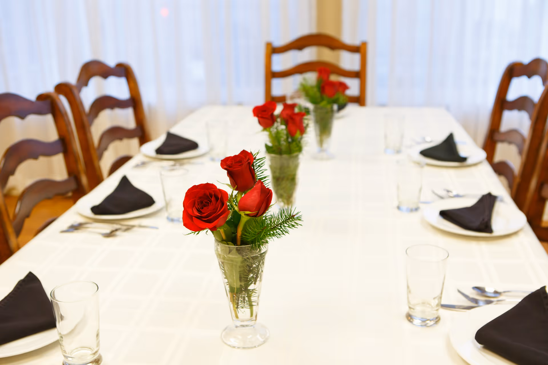 A dining table set with white tablecloth, black folded napkins on white plates, clear drinking glasses, and silverware. The table is decorated with small glass vases containing red roses and green foliage. Wooden chairs surround the table and sheer white curtains are visible in the background.