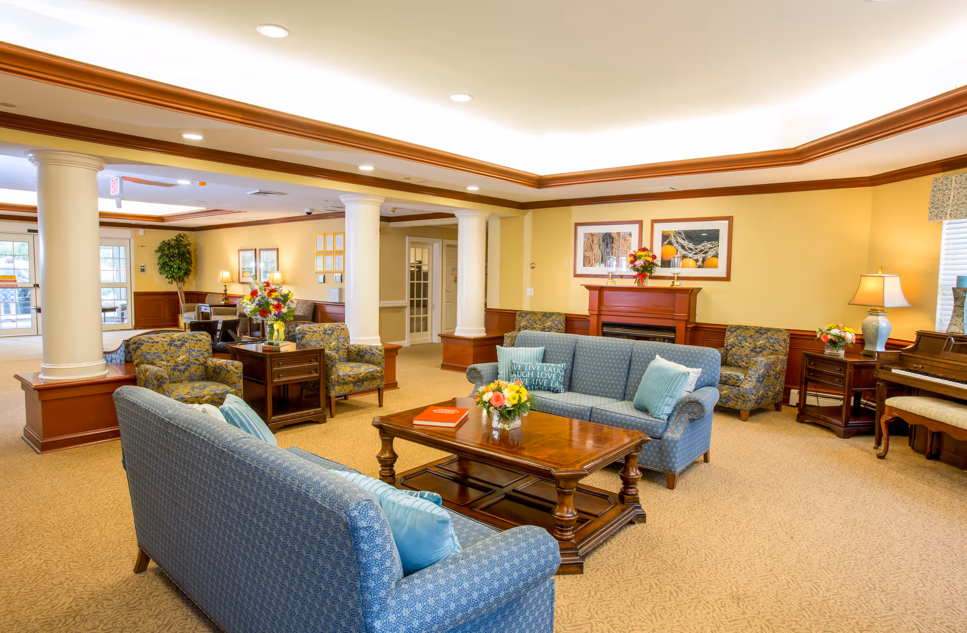 A spacious and well-lit living room area in a senior living facility with blue upholstered sofas and armchairs arranged around a wooden coffee table. The room features beige walls, a fireplace with framed artwork above it, decorative columns, and a piano in the corner. There are also table lamps, floral arrangements, and large windows allowing natural light to enter.