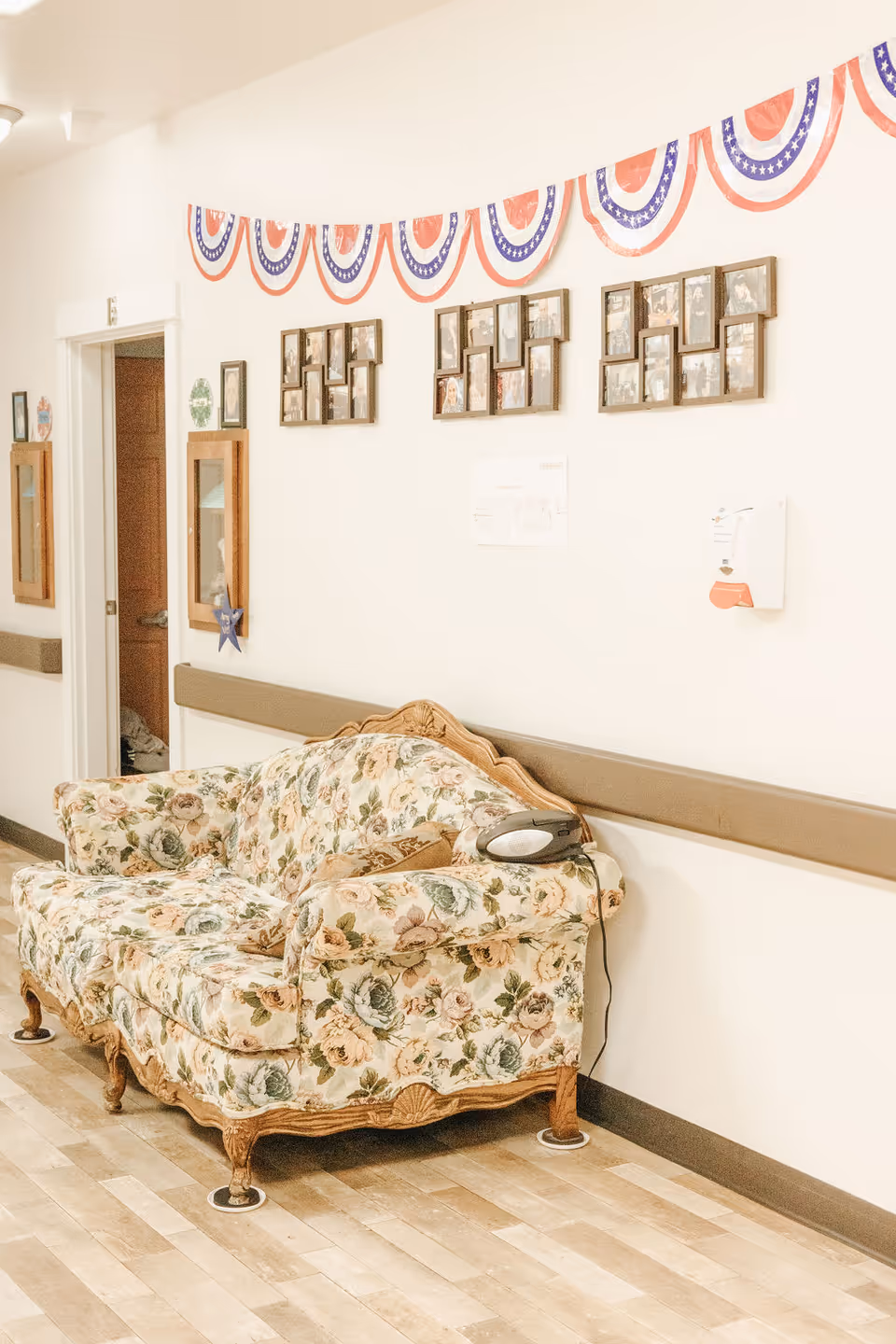 A floral patterned vintage sofa placed against a white wall in a hallway. Above the sofa, there are three photo collage frames and patriotic red, white, and blue bunting decorations. A hand sanitizer dispenser is mounted on the wall to the right of the sofa. The floor is wooden, and there is an open doorway visible on the left side of the image.