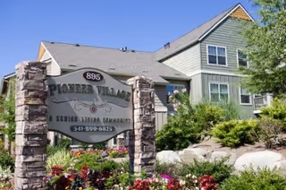 Exterior view of a senior living facility named Pioneer Village, showing a large sign with the address 895 and phone number 541-899-6825, surrounded by landscaped greenery and flowers, with a two-story building in the background under a clear blue sky.