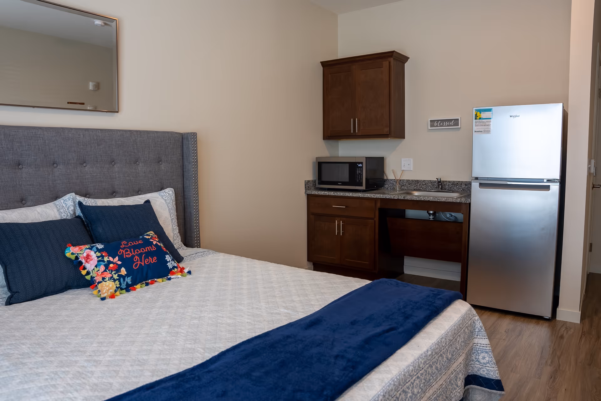 A senior living facility room featuring a neatly made bed with a gray upholstered headboard, blue and floral decorative pillows, and a blue throw blanket. To the right, there is a small kitchenette area with a granite countertop, a microwave, a stainless steel refrigerator, a sink, and wooden cabinets. A mirror hangs on the wall above the bed.