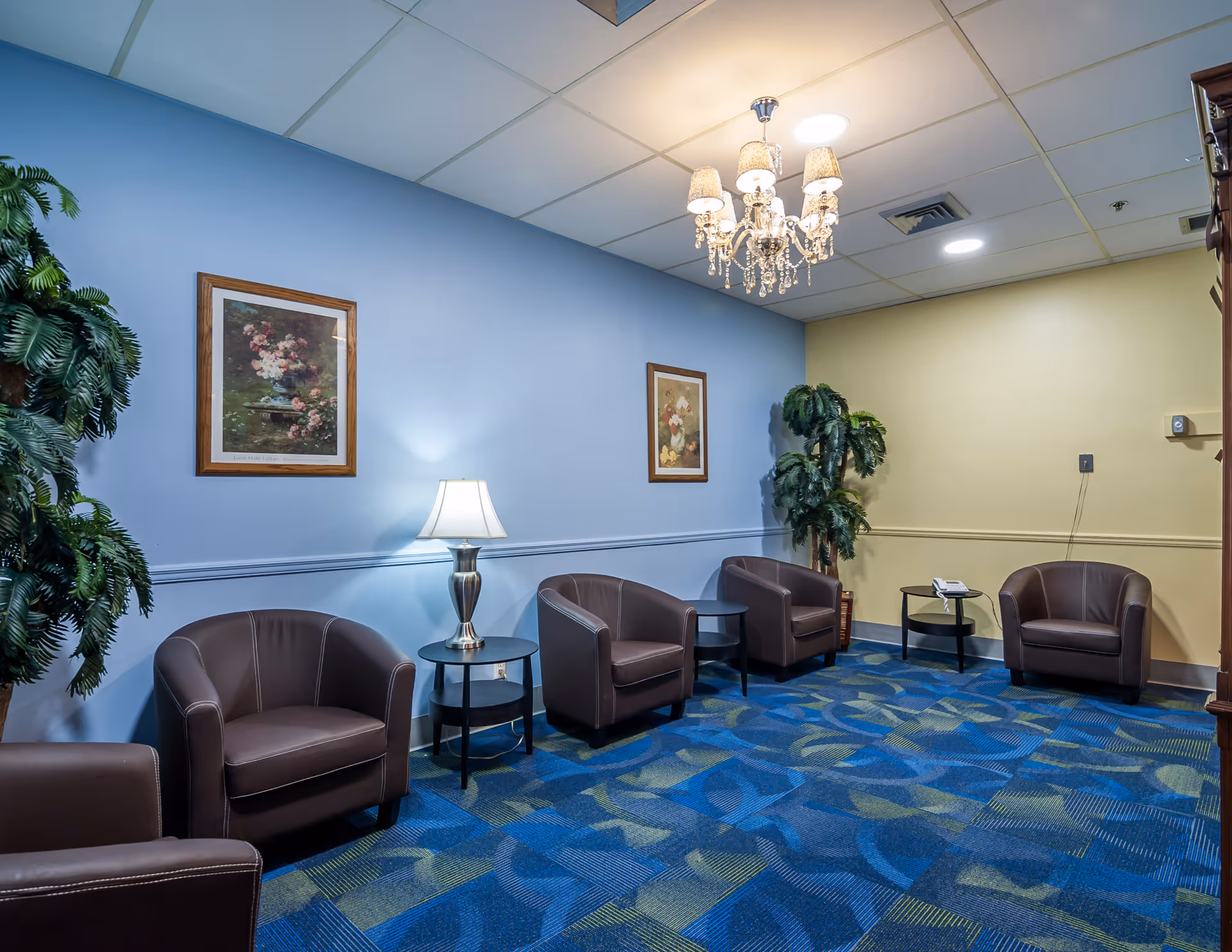 A cozy seating area in a senior living facility with five brown leather armchairs arranged along the walls. There are two small round tables with a lamp and a telephone on them. The walls are painted blue and beige, decorated with framed floral paintings. Two artificial plants are placed in the corners, and a chandelier hangs from the ceiling. The floor is covered with a blue patterned carpet.