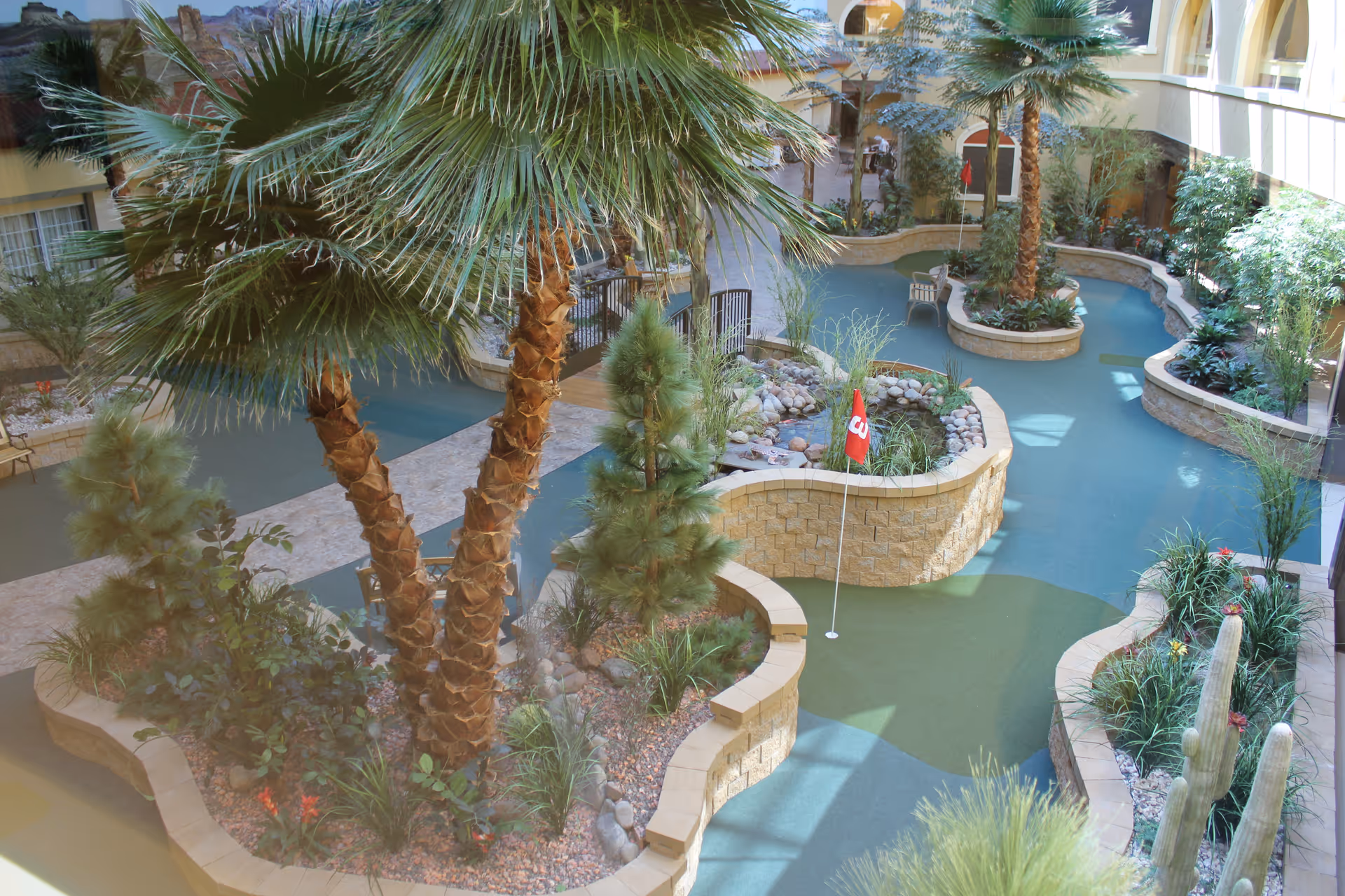 Indoor atrium with palm trees and a winding putting green surrounded by raised stone planters and landscaping.