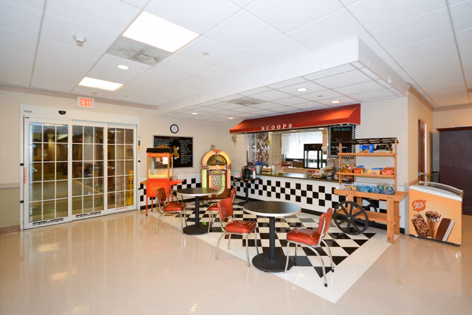 Interior view of a small retro-style snack bar area named 'SCOOPS' with black and white checkered tiles on the counter and floor. There are two round tables with red chairs, a popcorn machine, a jukebox, and a freezer with ice cream advertisements. Sliding glass doors are visible on the left side.