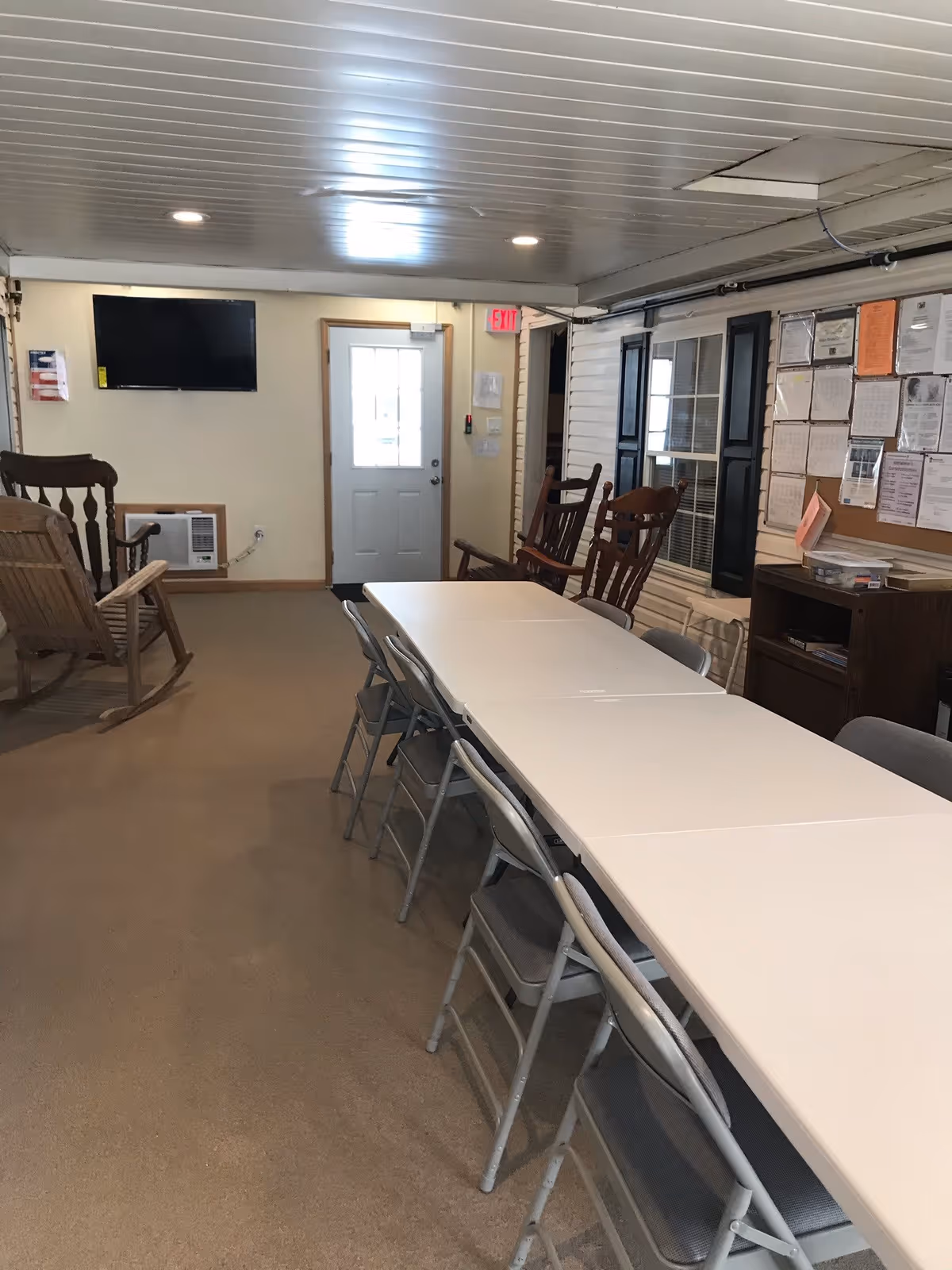 Interior room with a long white folding table surrounded by metal folding chairs. There are wooden rocking chairs along the walls, a wall-mounted TV, a door with a window, and a bulletin board with various papers pinned on it.