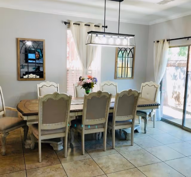 A bright dining room with a long wooden dining table surrounded by eight upholstered chairs. The room has tiled floors, two windows with white curtains, and a sliding glass door leading outside. A rectangular chandelier with multiple lights hangs above the table, and a vase with flowers is placed in the center. Two decorative mirrors with arched windowpane designs are mounted on the walls.