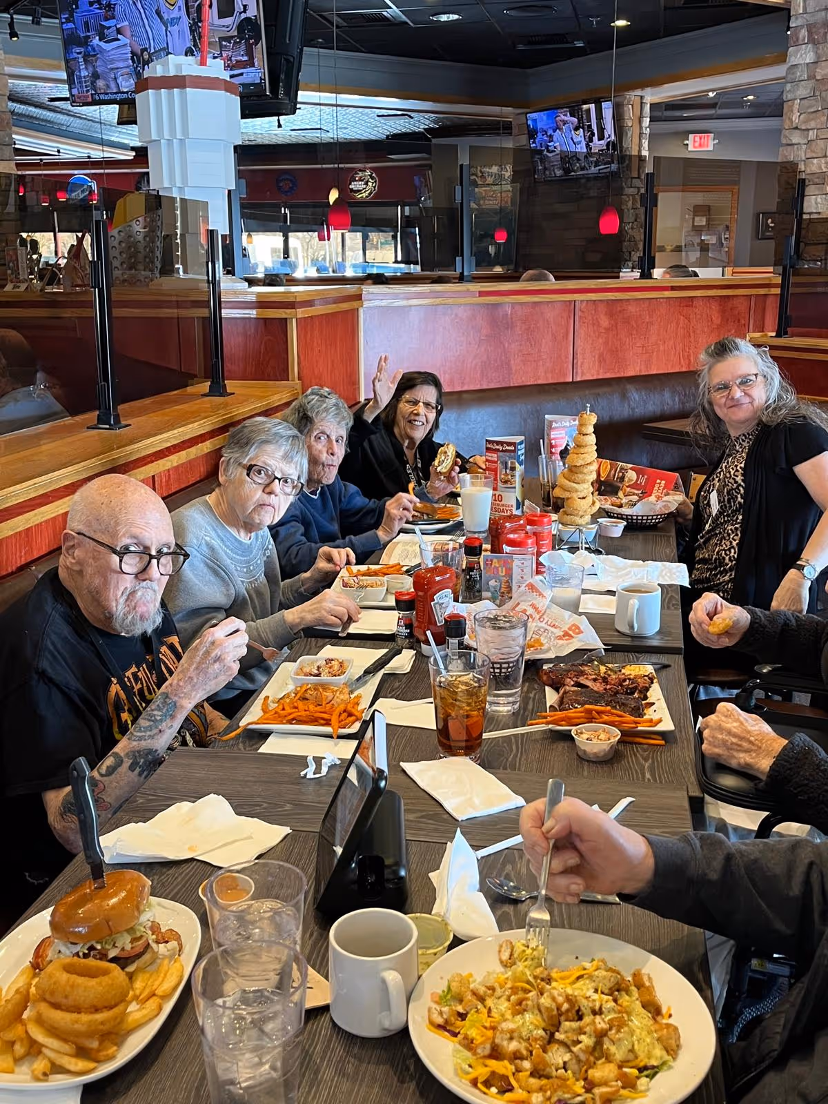 A group of elderly people sitting around a restaurant table enjoying a meal together. The table is filled with various dishes including burgers, onion rings, fries, and salads. The setting appears to be a casual dining restaurant with wooden paneling and multiple TV screens mounted on the walls.