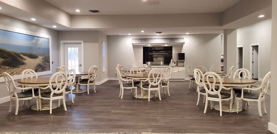 A spacious dining room with multiple round wooden tables surrounded by white chairs with circular backrests. The room has light-colored walls, a large framed beach scene on the left wall, and a door with a window. The floor is covered with dark wood-style flooring, and the ceiling has recessed lighting.