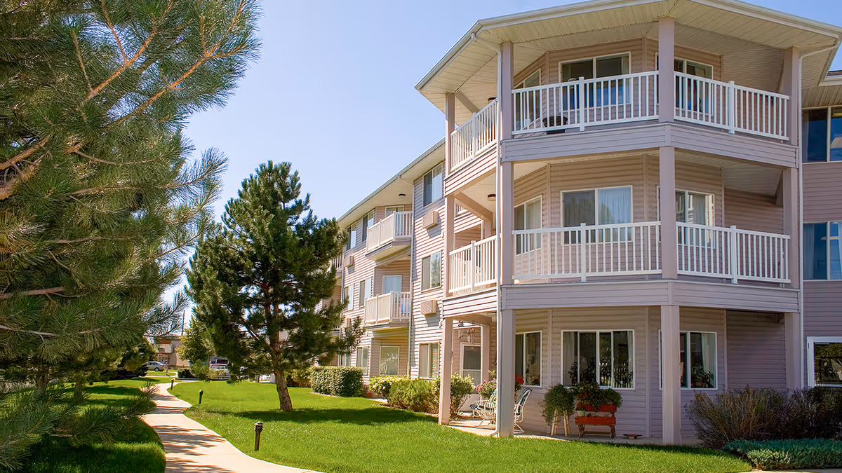 Exterior view of a multi-story residential building with balconies, surrounded by green grass, trees, and a paved walkway under a clear blue sky.