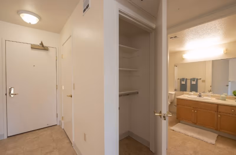 Interior view of a hallway in a senior living facility showing a white entrance door, a closet with open door revealing shelves and a hanging rod, and a bathroom with wooden cabinets, a large mirror, and a toilet visible in the background.