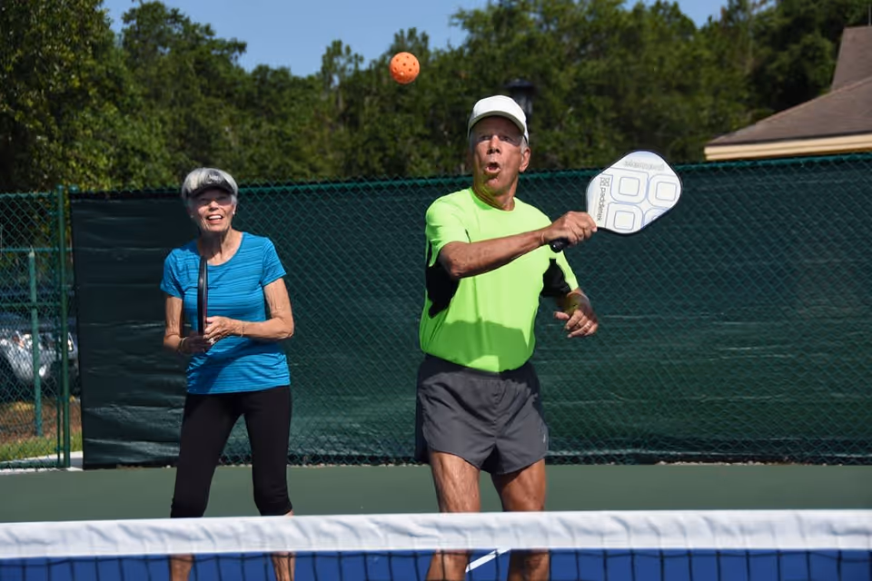 An elderly man and woman playing pickleball outdoors on a sunny day. The man, wearing a neon green shirt and gray shorts, is about to hit an orange pickleball with a paddle. The woman, dressed in a blue shirt and black pants, stands behind him holding a paddle and smiling. They are on a pickleball court with a green fence and trees in the background.