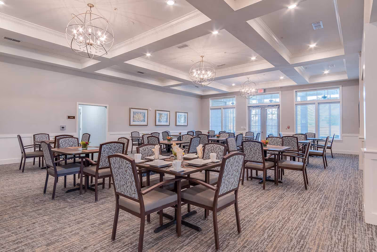Communal dining room with multiple set tables and upholstered chairs, decorative chandeliers, and large windows.