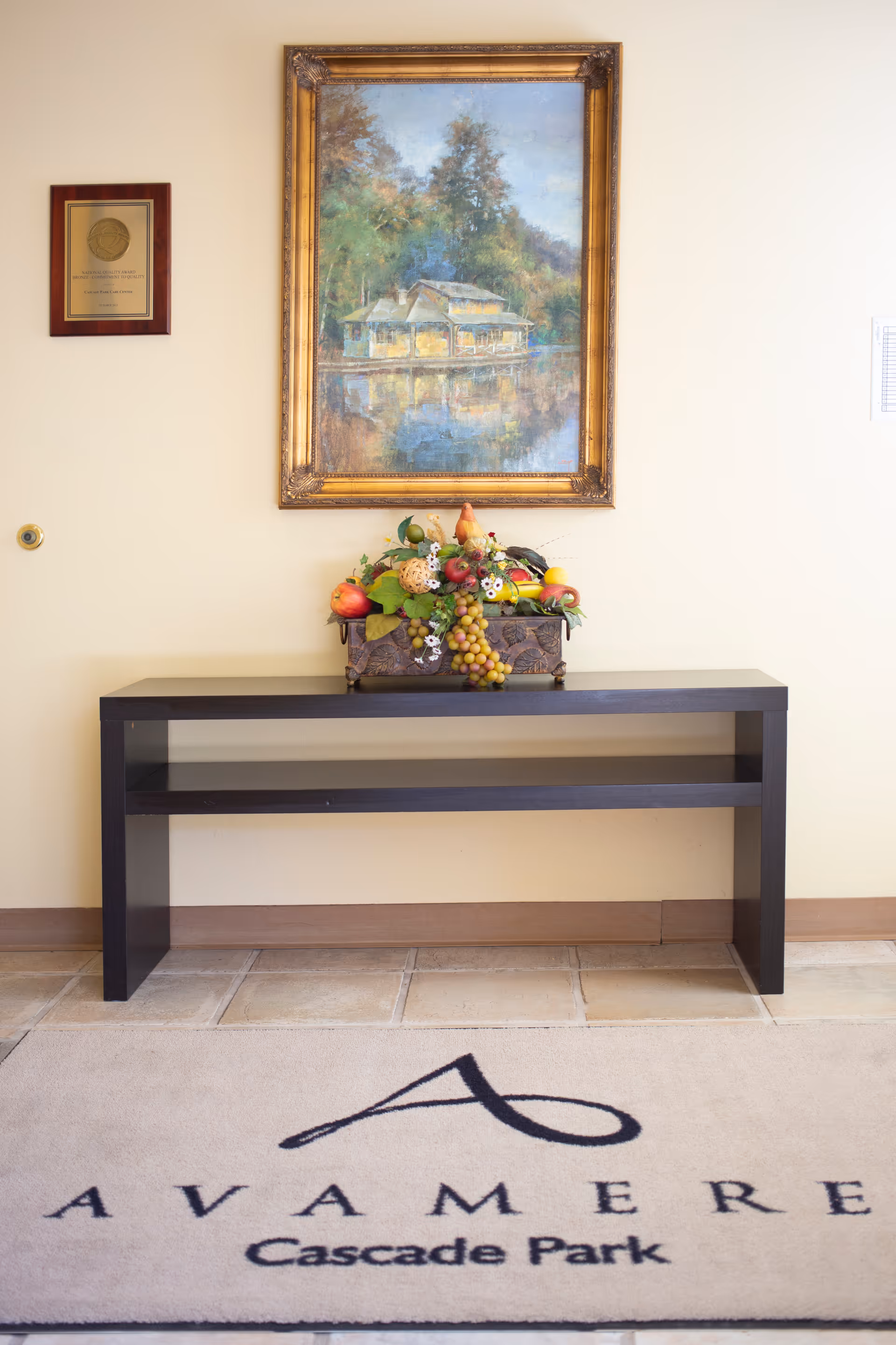 A black console table against a beige wall with a decorative fruit arrangement on top. Above the table hangs a framed painting of a house by a lake surrounded by trees. To the left of the painting is a plaque mounted on the wall. Below the table is a beige rug with the text 'AVAMERE Cascade Park' and a stylized letter 'A'.