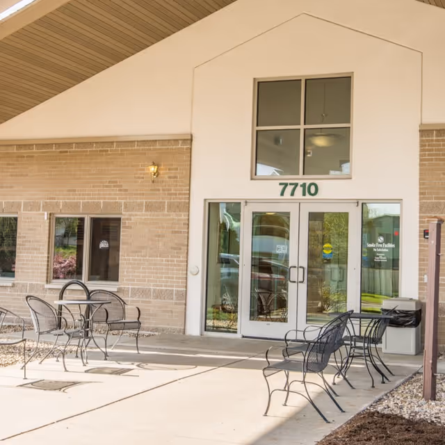 Entrance to a building with the number 7710 above double glass doors. There are metal outdoor tables and chairs on a concrete patio in front of the entrance. The building has a beige brick exterior with a light fixture on the wall and a trash can near the door.