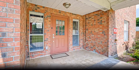 Covered entrance area of a brick building with a red door and two windows on either side. There are signs posted on the windows and a small mat in front of the door. The floor is tiled and there is some greenery visible on the right side.
