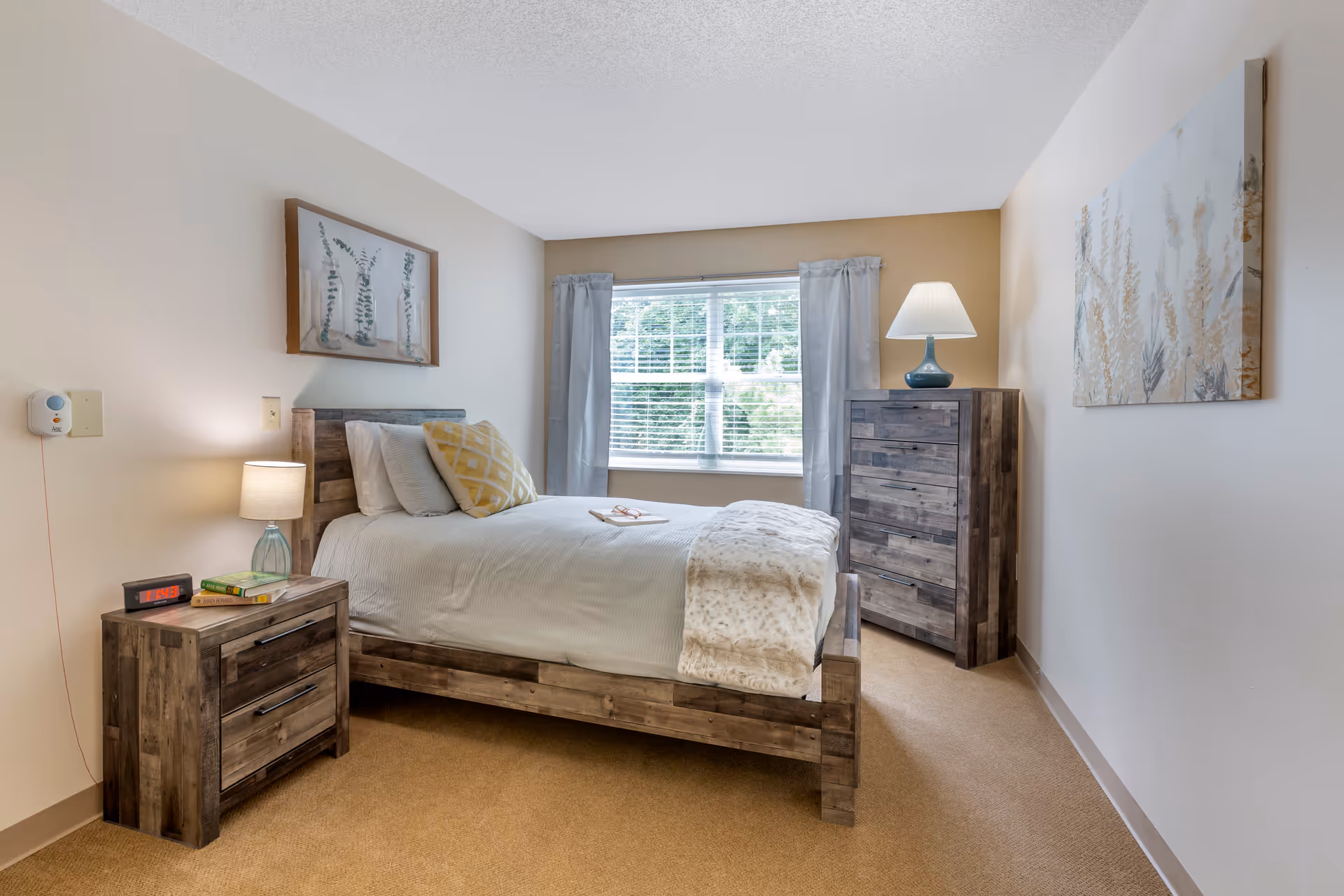 A cozy bedroom with a wooden bed frame, white bedding, and a yellow patterned pillow. There is a wooden nightstand with a lamp, books, and an alarm clock. A window with gray curtains lets in natural light. A wooden dresser with a lamp on top is against the far wall, and two pieces of wall art decorate the room.