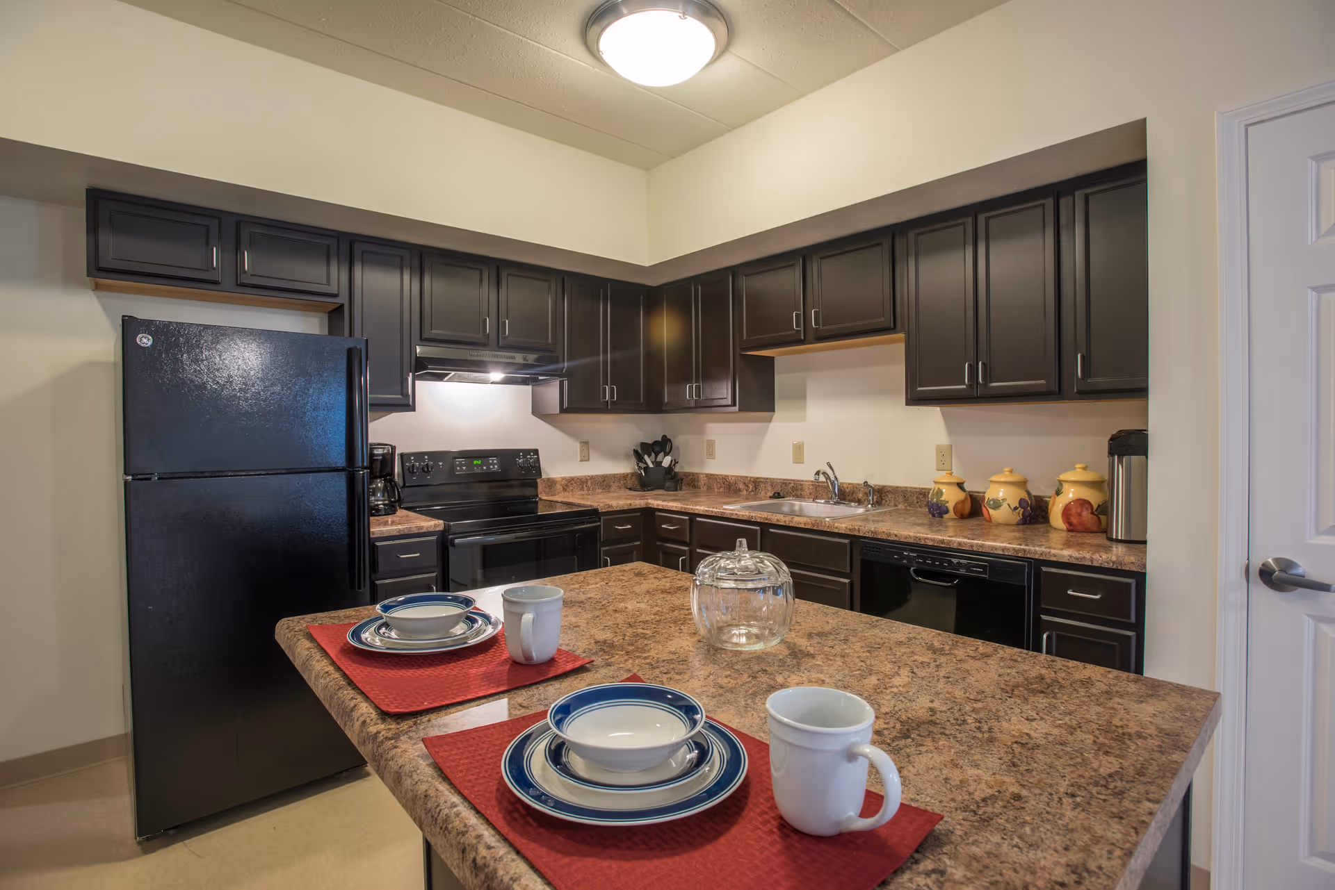 A modern kitchen with dark brown cabinets, a black refrigerator, stove, and dishwasher. The countertop is a brown marble pattern with two place settings including plates, bowls, and mugs on red placemats. There are decorative jars and a glass container on the counter.