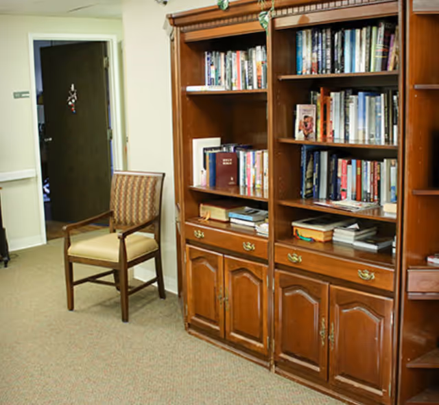 Interior view of a room with a wooden bookshelf filled with books and a wooden chair with a cushioned seat and backrest. There is a door in the background and beige carpeted flooring.
