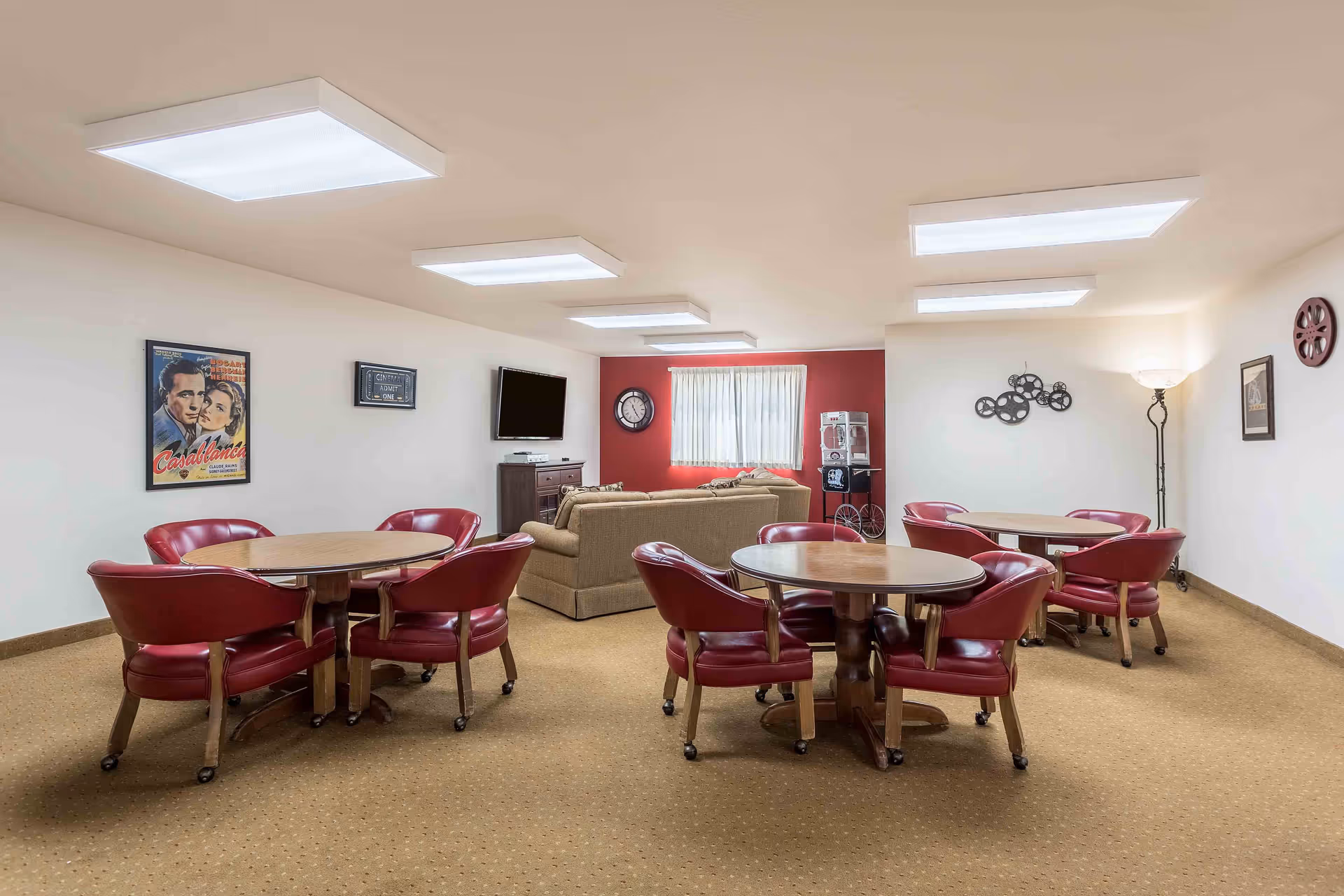 A common area with three round wooden tables each surrounded by four red cushioned chairs on wheels. In the background, there is a beige sofa facing a wall-mounted flat screen TV. The walls are decorated with movie-themed art including a Casablanca poster, a clock, and film reel decorations. A popcorn machine is placed near the window with white curtains and a red accent wall behind it. The room has beige carpeting and bright ceiling lights.