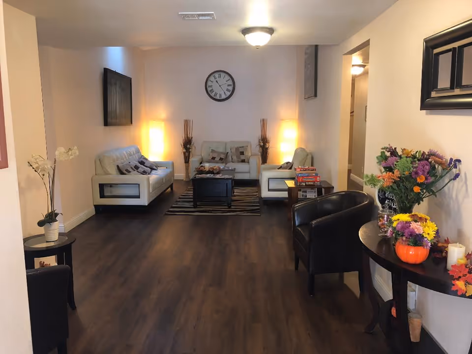 A cozy living room area with three white sofas arranged around a black coffee table on a striped rug. Two tall lamps and decorative vases with dried plants are placed against the back wall, which also features a large clock. The room has dark wood flooring and light-colored walls. On the right side, there is a small round table with colorful flower arrangements and a black armchair nearby.