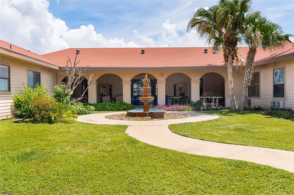Sunlit courtyard with a central tiered fountain, curved walkway, palm trees and a single-story building with an arched colonnade.