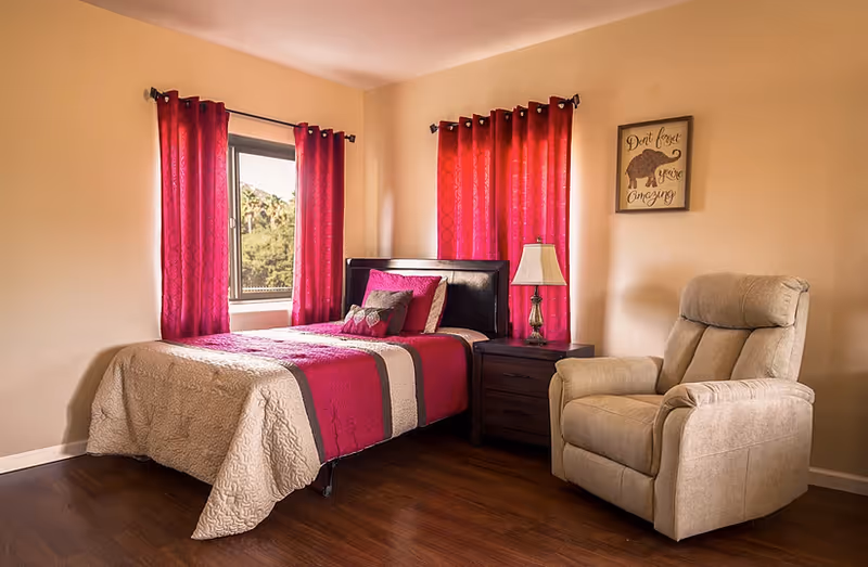 A cozy bedroom with a single bed dressed in pink and beige bedding, red curtains, a nightstand with a lamp, and a beige recliner chair.