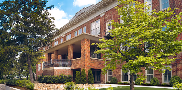 Exterior view of a multi-story brick senior living facility with large windows, a covered balcony, and surrounded by green trees and shrubs under a partly cloudy sky.