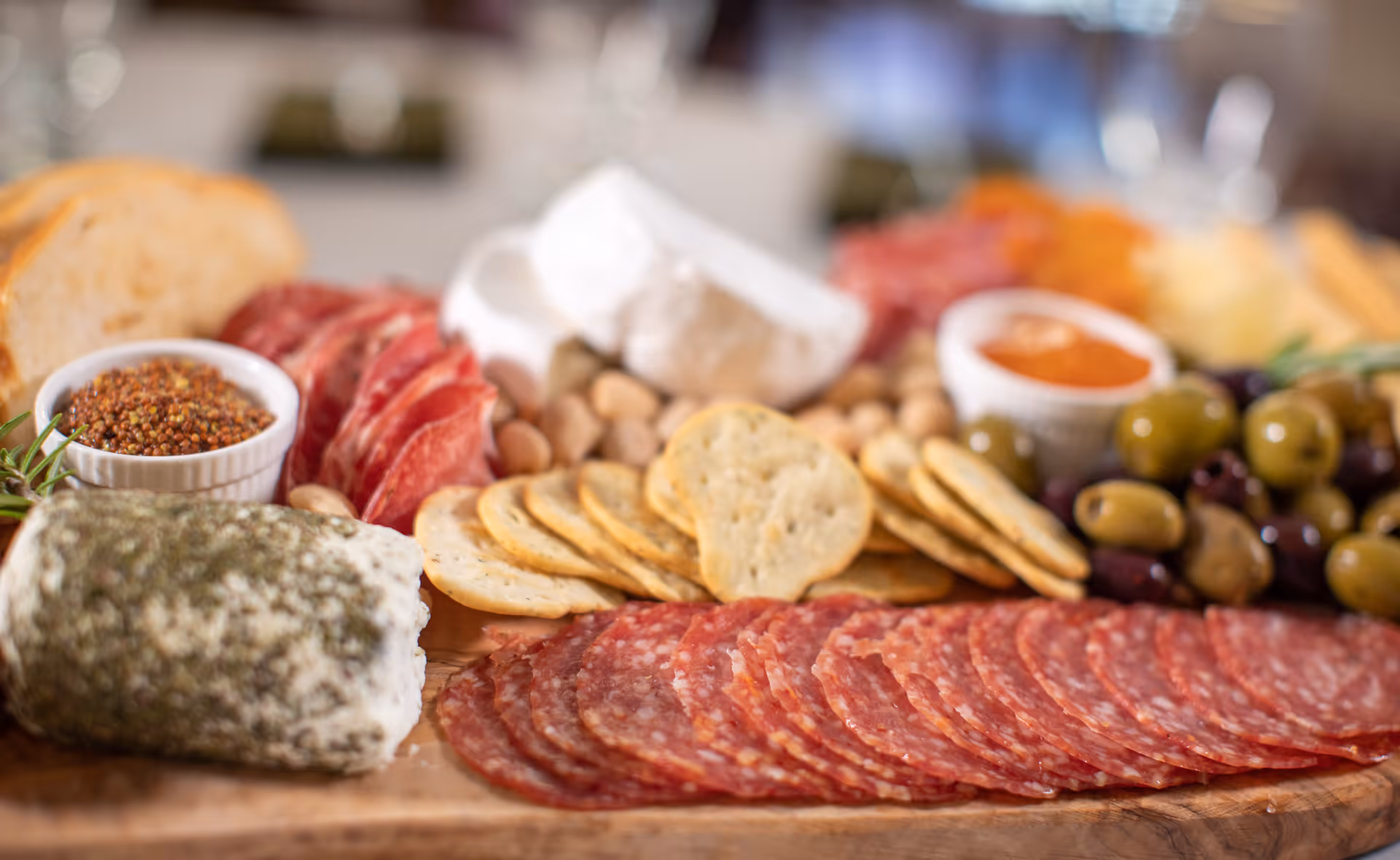Close-up of a charcuterie board featuring slices of salami, crackers, green and black olives, a log of herb-coated cheese, a wedge of soft white cheese, slices of bread, and small bowls of mustard and red sauce.
