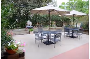 Outdoor patio area with metal tables and chairs under large beige umbrellas, surrounded by greenery and potted plants.