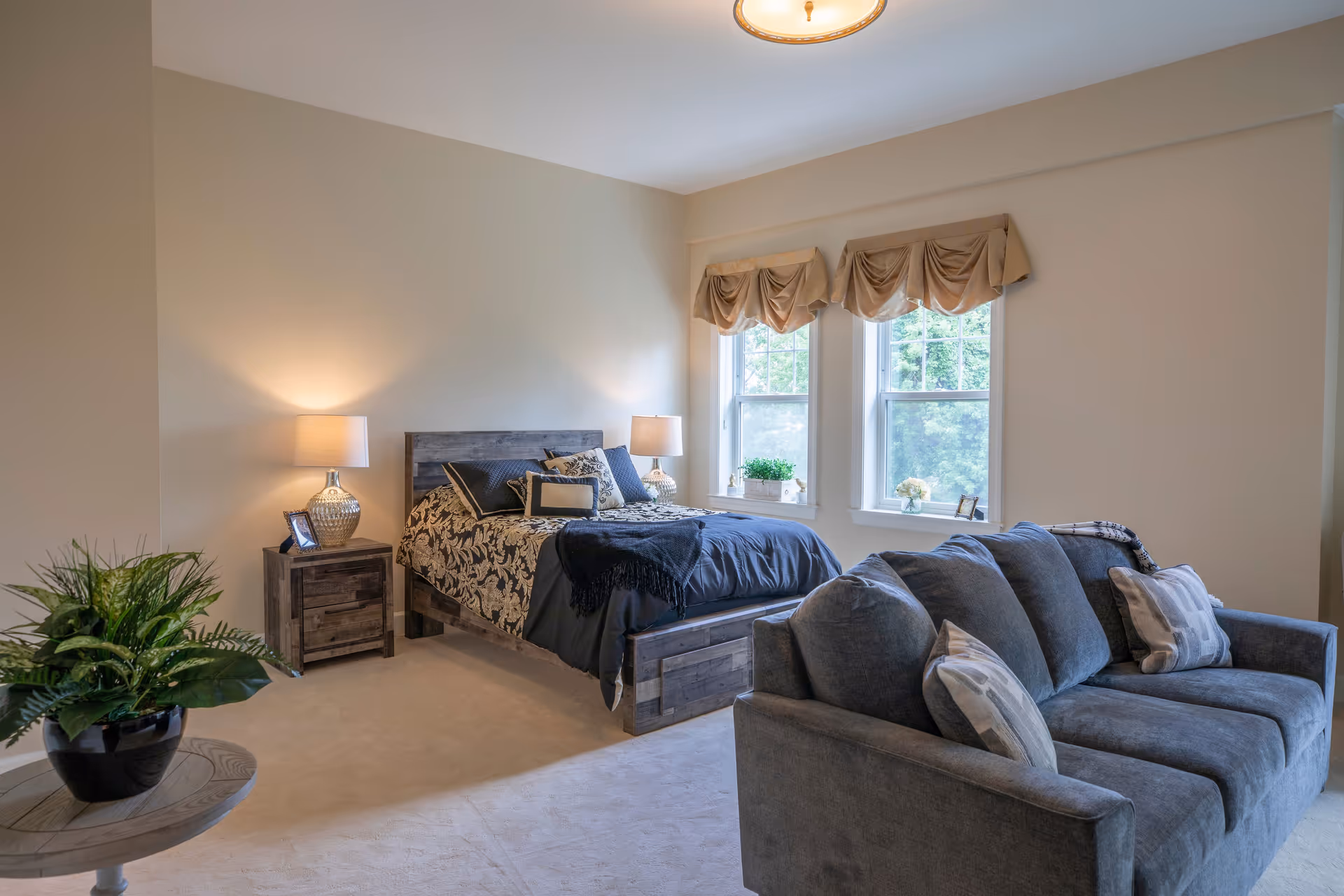 A cozy bedroom with a wooden bed featuring a dark patterned bedspread and multiple pillows. Two bedside tables with lamps flank the bed. There are two windows with beige valances letting in natural light. A gray sofa with cushions is positioned in front of the bed, and a round table with a potted plant is in the foreground.