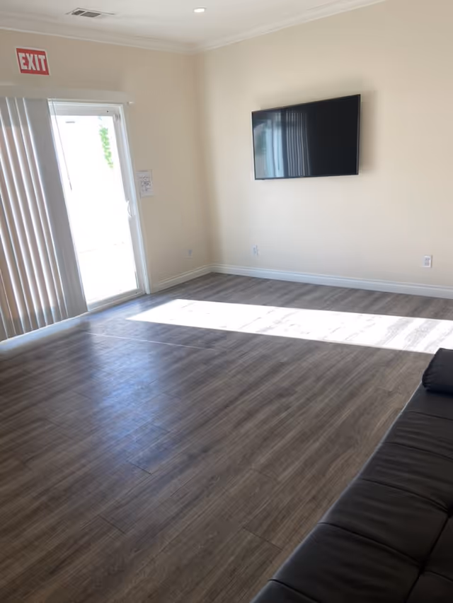 A bright room with wood flooring, a wall-mounted flat screen TV, a black leather couch partially visible on the right, and a glass door with vertical blinds leading outside. An exit sign is mounted above the door.