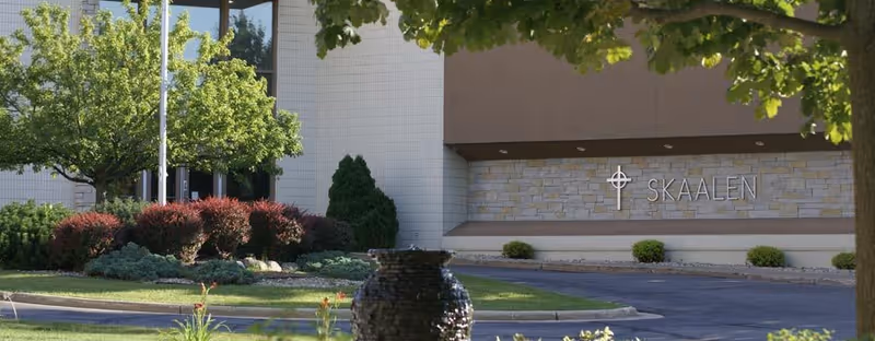 Exterior view of Skaalen Retirement Services building with landscaped greenery including trees, bushes, and a stone water feature in the foreground.