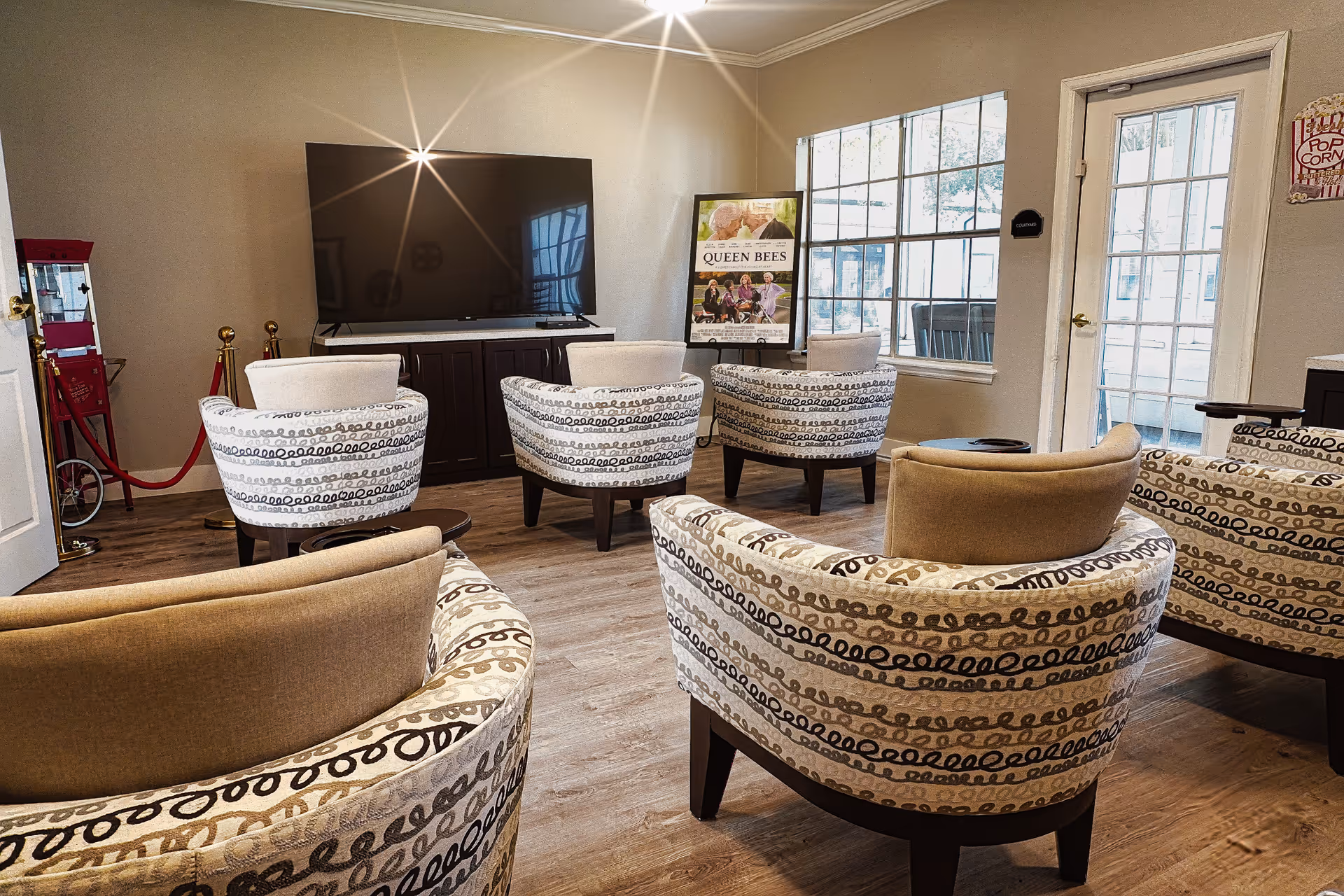 A cozy common area with patterned armchairs arranged in rows facing a large flat-screen TV mounted on a cabinet. There is a popcorn machine on the left side and a poster stand near a window and glass door that leads outside. The room has wooden flooring and neutral-colored walls.
