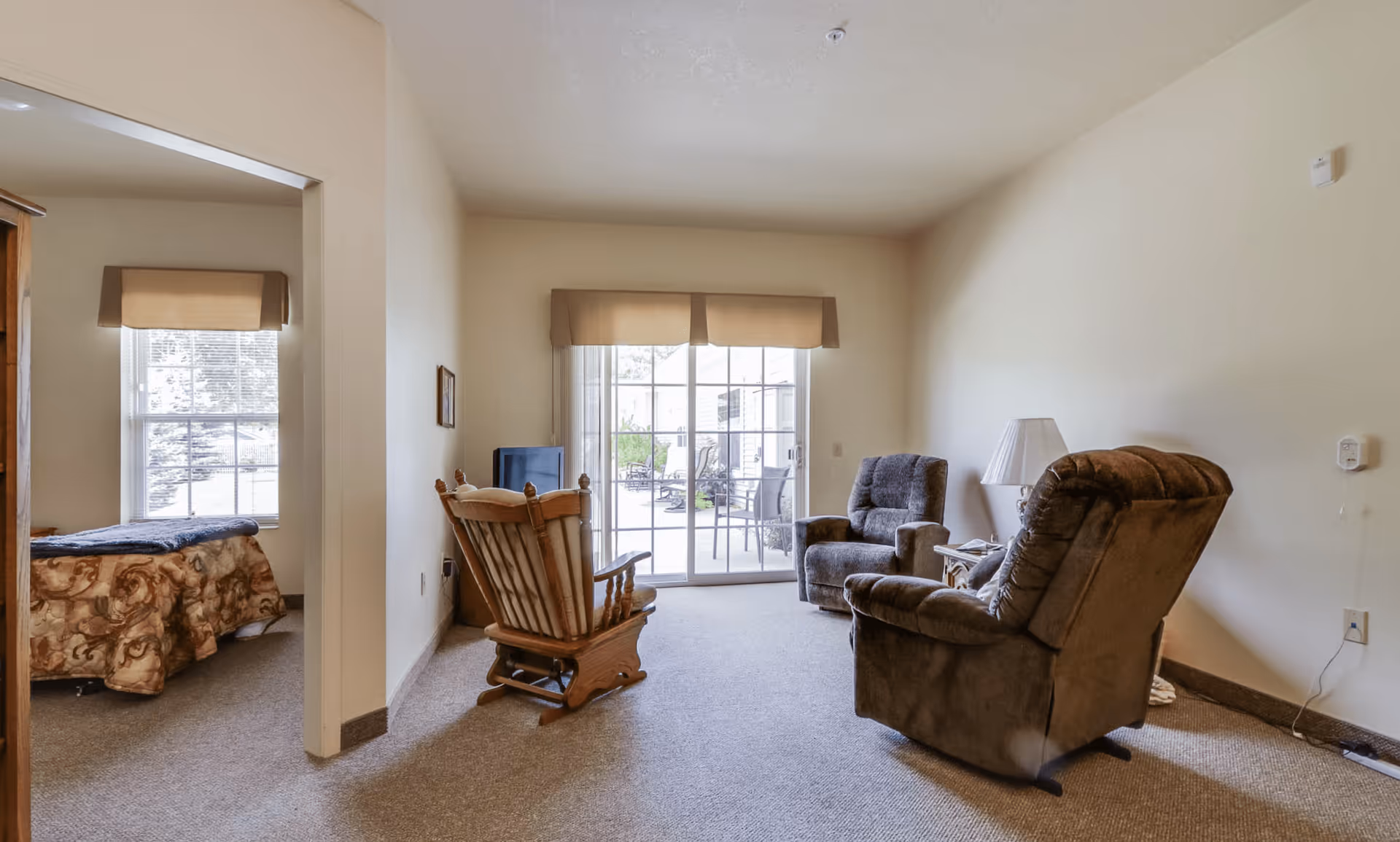 A cozy living room with a sliding glass door leading to an outdoor patio. The room contains a wooden rocking chair, two cushioned armchairs, a small side table with a lamp, and a television on a stand. To the left, an open doorway reveals a bedroom with a window and a bed covered with a patterned blanket.