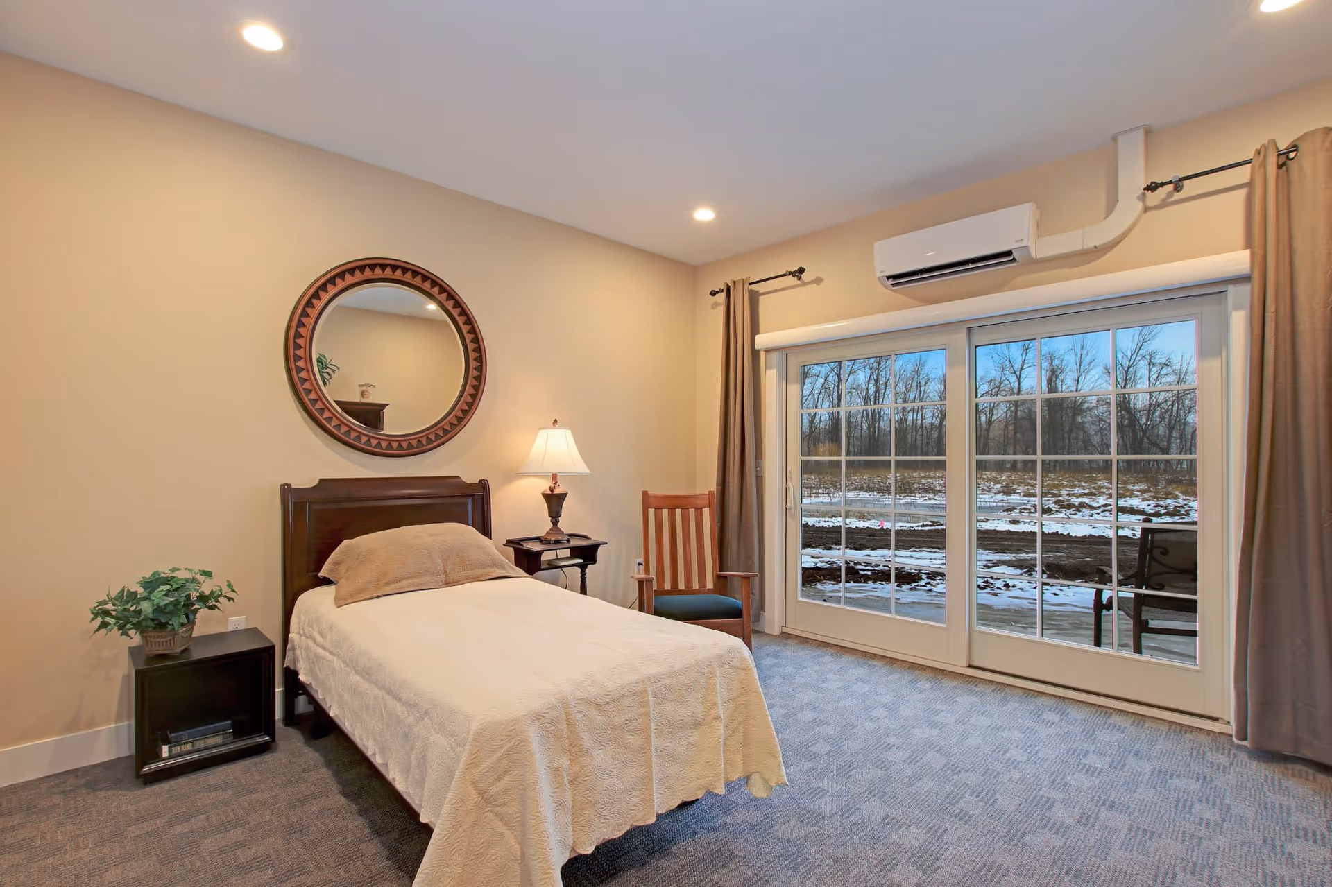 A bedroom with a single bed covered with a beige blanket and a brown pillow. There is a wooden headboard, a nightstand with a lamp and a small plant, a wooden chair, and a large sliding glass door with curtains showing a snowy outdoor scene. A round decorative mirror hangs on the wall above the bed.