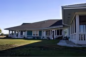 Single-story senior living building with covered porches, a walkway, and a grassy lawn under a clear blue sky.