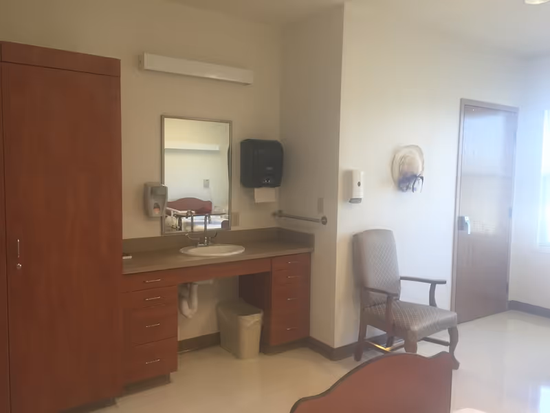 A room with a wooden cabinet, a countertop with a sink, a mirror above the sink, a paper towel dispenser, a soap dispenser, a trash bin under the sink, a cushioned armchair, a closed wooden door, and a window letting in natural light.