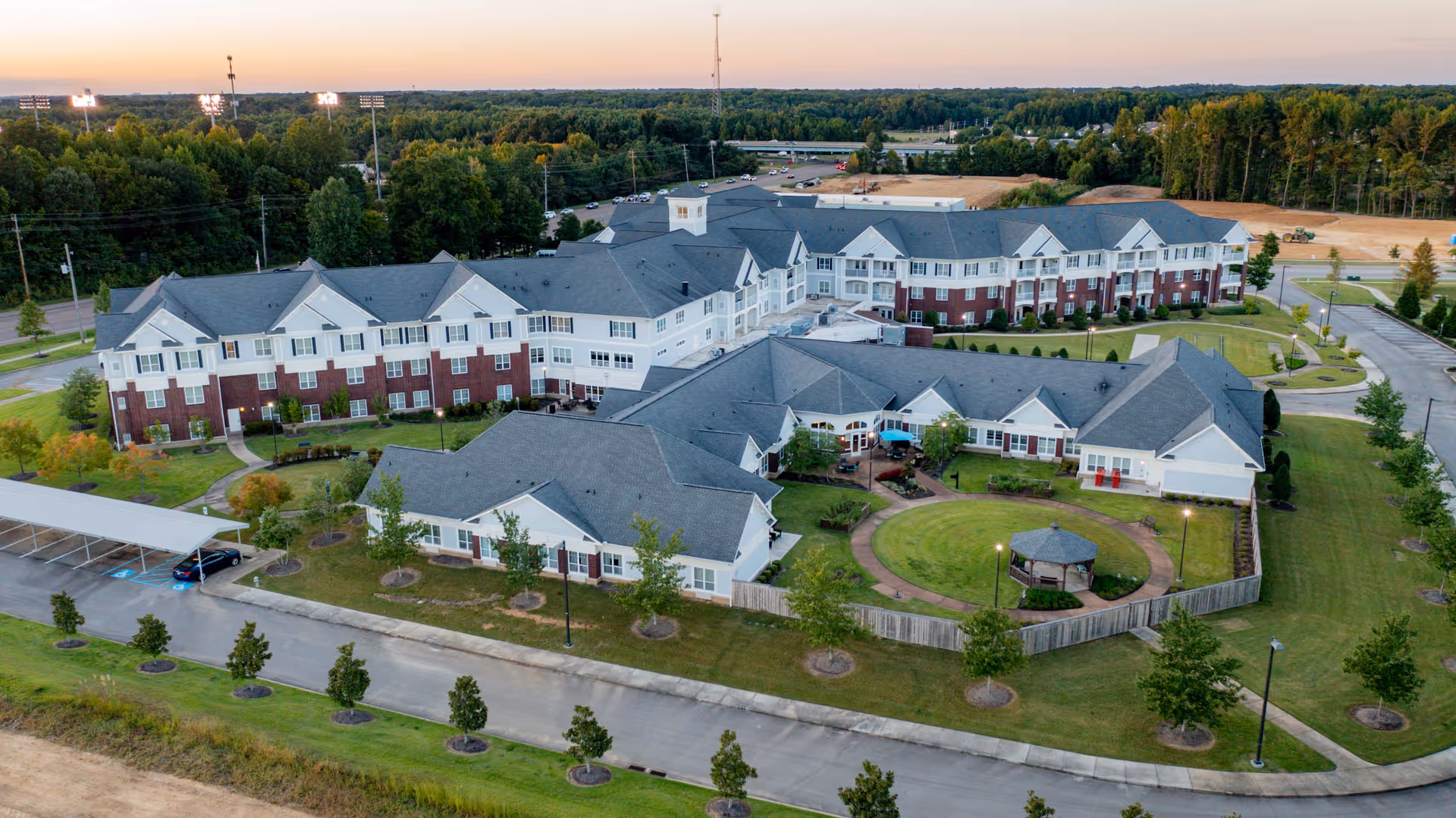 Aerial view of a multi-wing assisted living facility with a central circular courtyard and gazebo, surrounding parking, and tree-lined grounds.