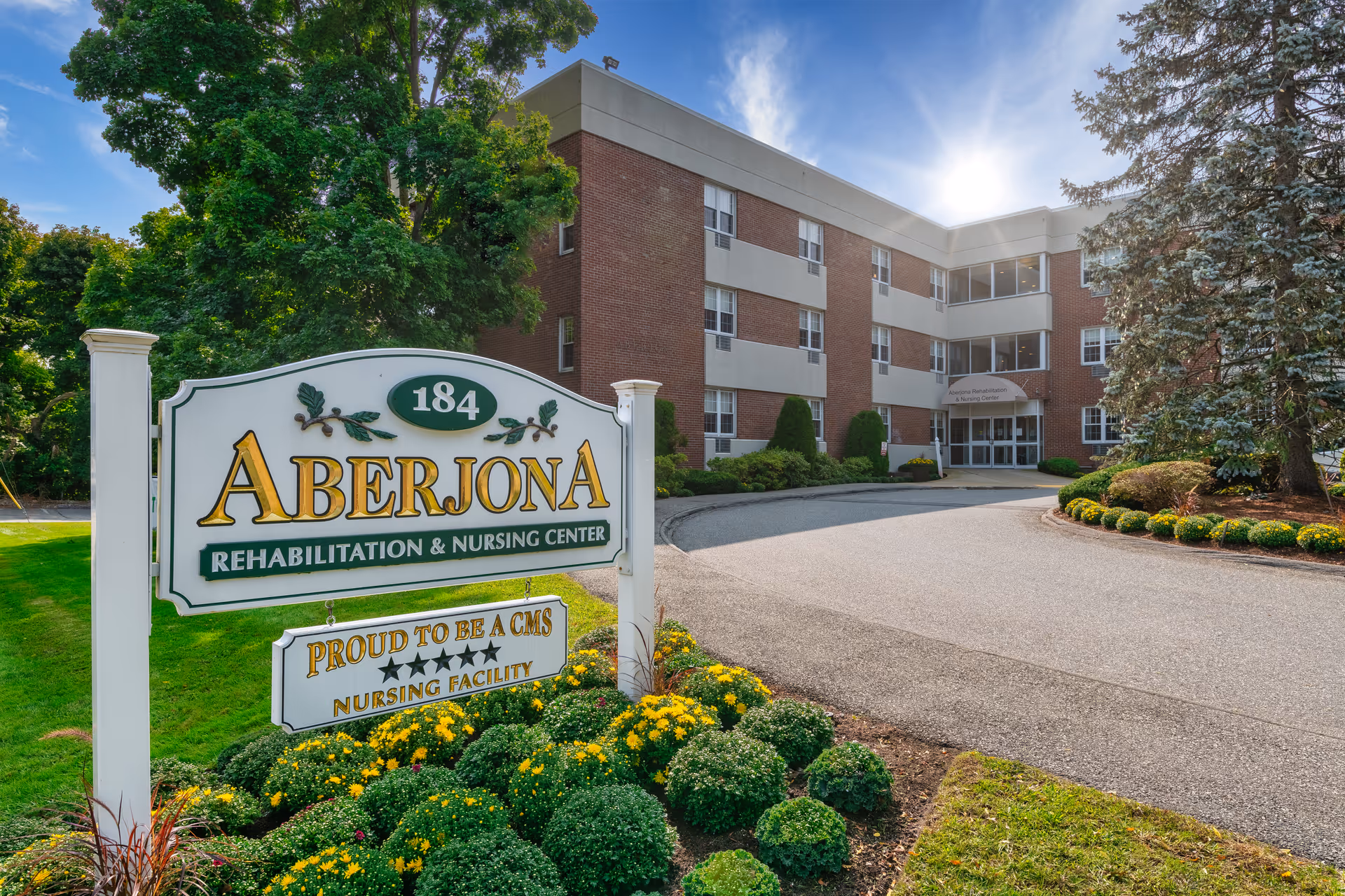 Exterior view of Aberjona Rehabilitation & Nursing Center building with a large sign in the foreground displaying the facility name and a note about being a 5-star CMS nursing facility. The building is a three-story brick structure with multiple windows, surrounded by trees, shrubs, and a curved driveway leading to the entrance.